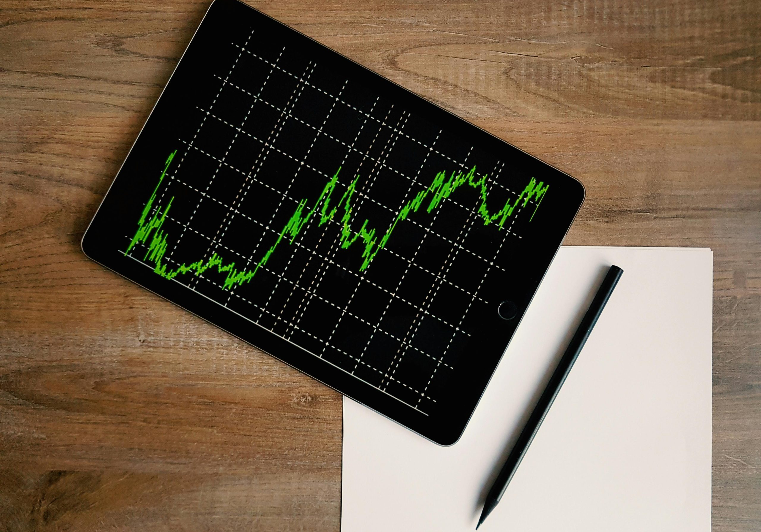 Flatlay of an iPad displaying stock market graph on a wooden desk with a pencil and paper.